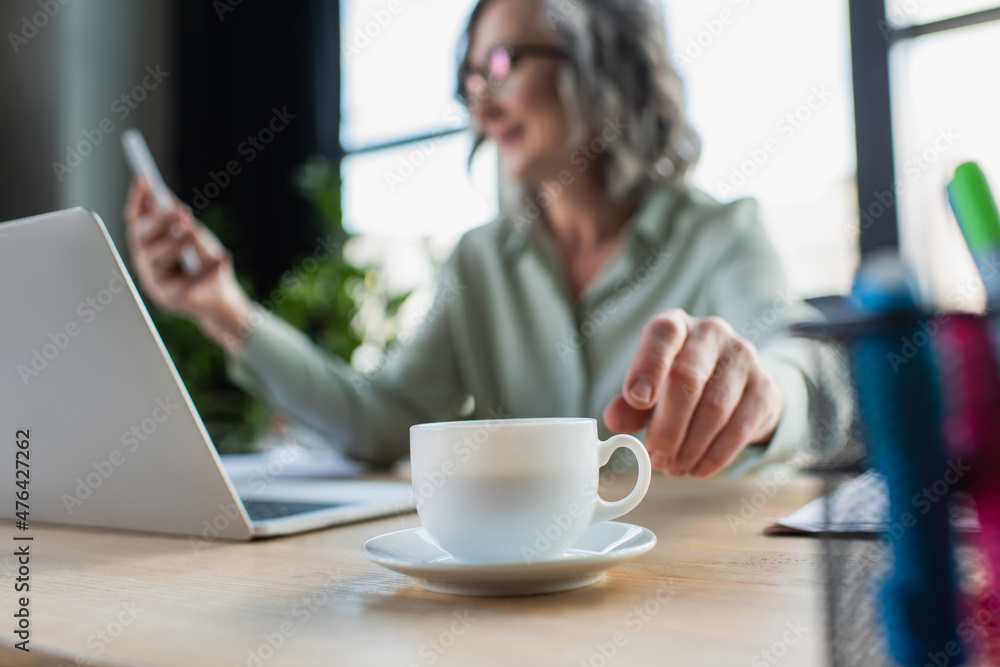 Cup of coffee near blurred businesswoman and laptop in office. Cup of coffee near blurred businesswoman and laptop in office.
