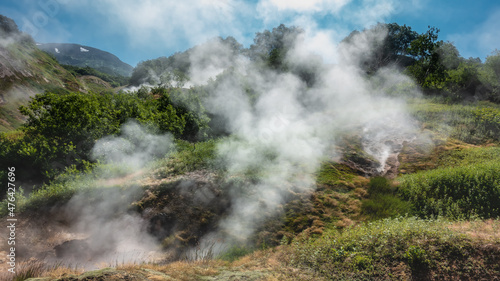 Thick smoke and steam rise from the fumaroles . There is green vegetation all around. Mountains against the blue sky. Valley of Geysers. Kamchatka