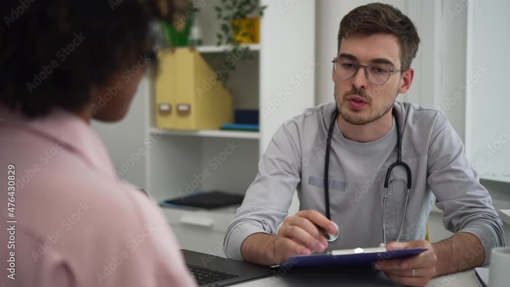 Doctor and patient at consultation. Handsome young male therapist in grey uniform explains spbd treatment protocol to black lady in hospital office Doctor and patient at consultation. Handsome young male therapist in grey uniform explains spbd treatment protocol to black lady in hospital office