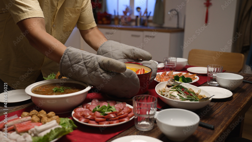 man wearing protective gloves carrying and serving hot soup in pot on the dining table, preparing for Chinese new year dinner at home