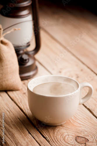 Cup with coffee with milk and hot smoke, on old wooden table, raffia sack and lamp.