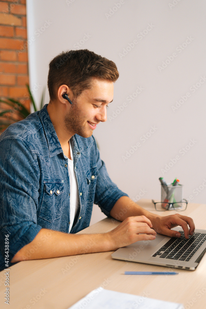 Vertical portrait of cheerful handsome freelance programmer male in wireless earphone working on laptop computer sitting at desk at home office. Concept of distance remote work, education. Vertical portrait of cheerful handsome freelance programmer male in wireless earphone working on laptop computer sitting at desk at home office. Concept of distance remote work, education.