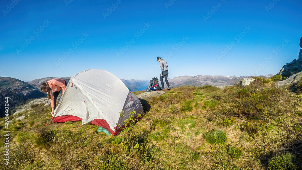 Fototapeta premium A young couple putting the tent up in the wilderness. The girl struggles with the strings,the boy brings the things from a big hiking backpack in the back. Camping in the nature. Bright and sunny day
