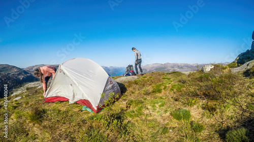 A young couple putting the tent up in the wilderness. The girl struggles with the strings,the boy brings the things from a big hiking backpack in the back. Camping in the nature. Bright and sunny day photo