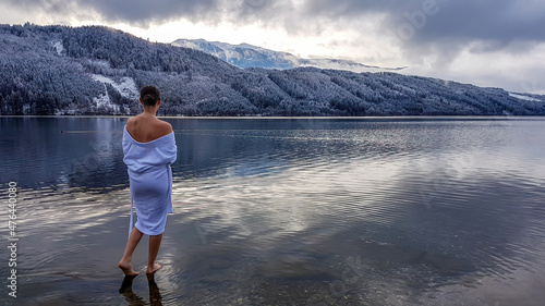 A girl in a white bathrobe walking in a shallow water of a lake. Half of the back of a girl is naked. On the other side of the lake there are mountains covered with snow. Winter alpine spa. photo