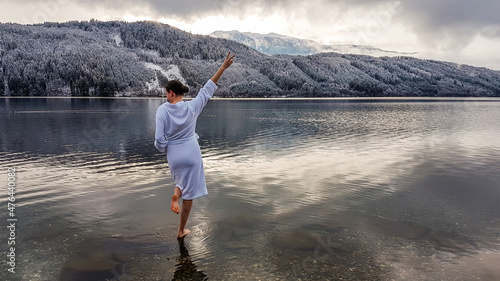 Girl in a white bathrobe walking in a shallow water of a lake. Girl is holding one hand up  gesture of happiness. On the other side of the lake there are mountains covered with snow Winter alpine spa