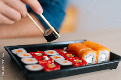 Close-up front view hands of unrecognizable young man holding sushi with chopsticks sitting at desk. Closeup of male enjoying sushi at workplace, selective focus, blurred background.