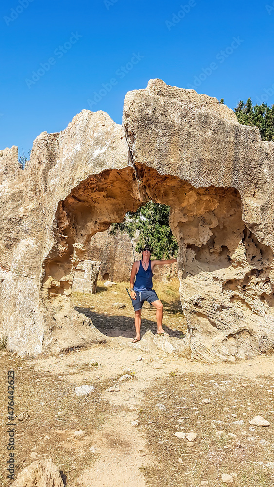 A young man wandering around Kato Paphos Archeological Park. He is standing under an arch, which could have previously been part of a bigger building. Dry grass on the ground. Sandstone structure.