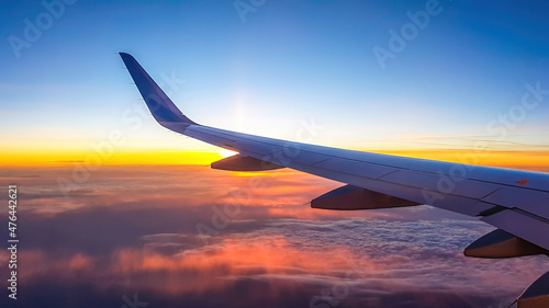 A colorful sunrise seen from the plane window. Sun is raising from the clouds, painting them pink and orange. Horizon line turns yellow with the sunbeams. Plane wing covering the sun. Romantic view