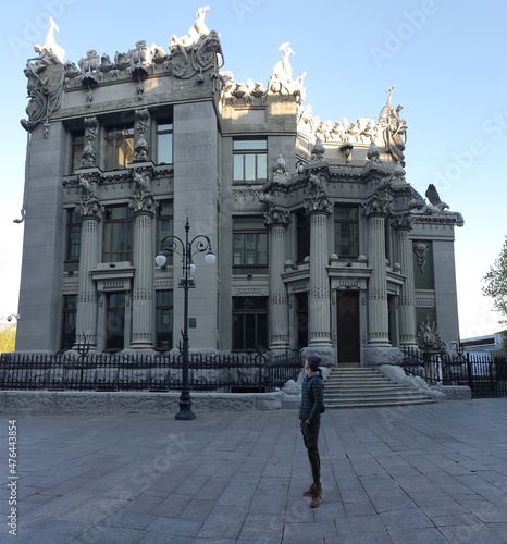 A young man in a green jacket standing in front of House with Chimaeras, Kiev, Ukraine. Sunlight is reaching the highest parts of the building. HOse is nicely decorated with different animals. photo