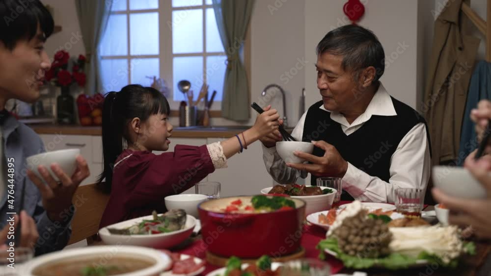 happy asian grandfather talking with hand gestures as his polite granddaughter putting food into his bowl at dinner table on chinese lunar new year's eve at home happy asian grandfather talking with hand gestures as his polite granddaughter putting food into his bowl at dinner table on chinese lunar new year's eve at home