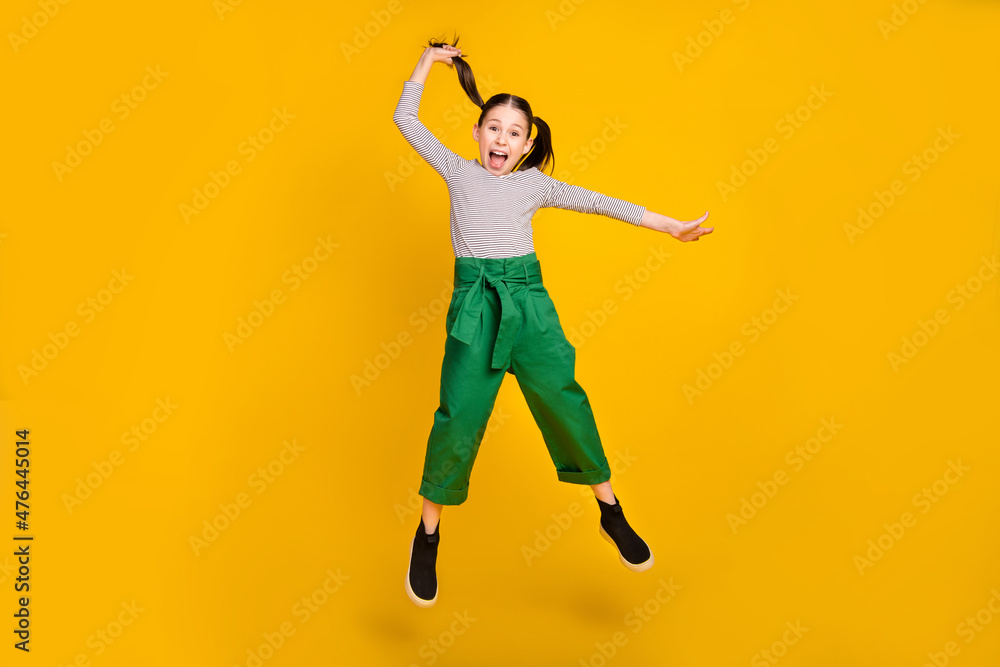 Full length photo of young excited school girl jump up hold ponytail isolated over yellow color background