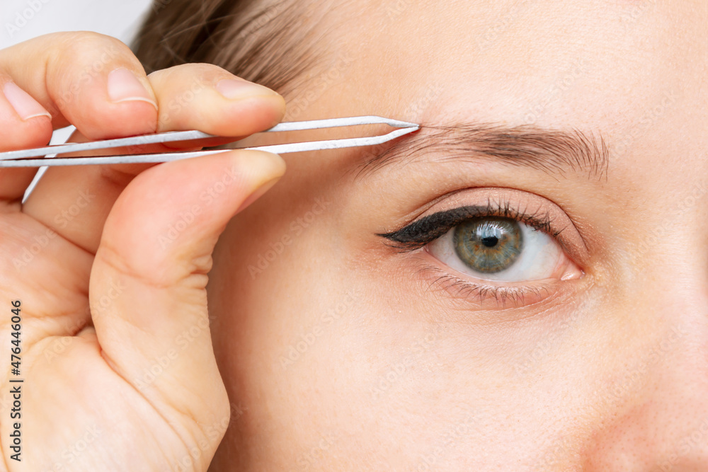 Fototapeta premium Close-up of the face of a young caucasian pretty woman plucking eyebrows with tweezers. Eyebrow shape correction. Cosmetology, beauty 