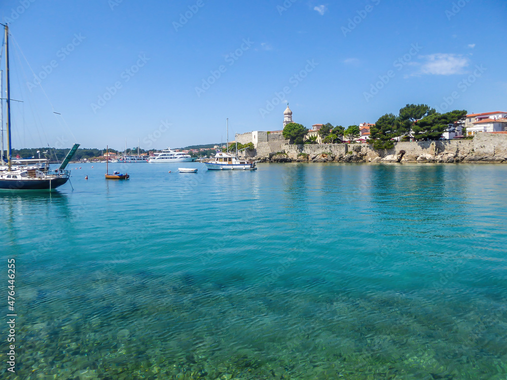 Sea view on Croatian city Krk. The city is build directly by the sea. Thick walls emerge high up from the sea line, defence building. There is a church tower, towering above other constructions.