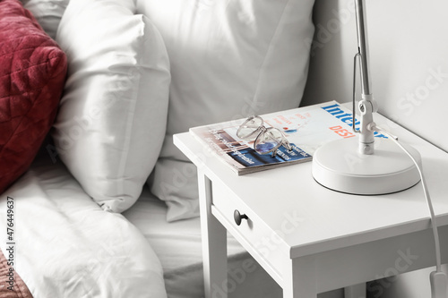 Bedside table with eyeglasses and magazine near light wall