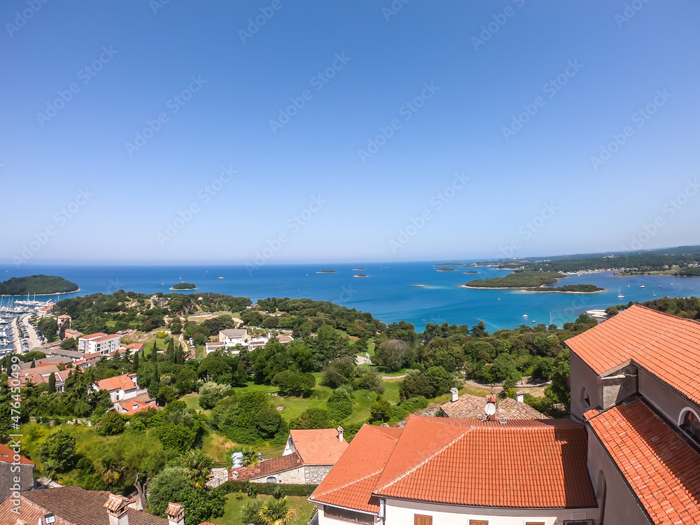 A remote view on the harbour from a hill. Rows of docks waiting for the boats and yachts to anchor there. Lots of houses surrounding the port. In the back there are few islands. Lush green plants