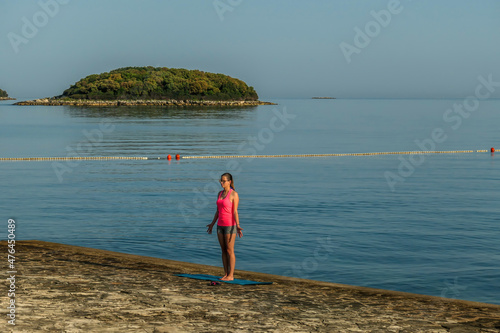 A girl in pink T-shirt practicing yoga by the seashore. She is standing in a mountain pose. There are few islands in the back. Clear and sunny day, calm sea. Outdoor practice healing mind and body.