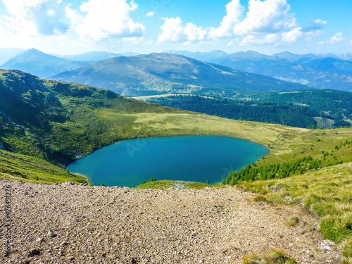 A view on an Alpine lake, hiding in between tall mountains. The lake has dark blue color, surrounded by lush green grass. Endless chains of mountains visible. There is viewing platform above the lake