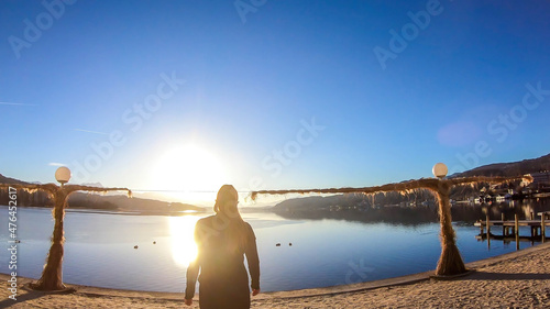 A girl in a black outfit walks along the lakeside. There is a scarecrow next to the shore. The surface of the lake is still and calm. There are tall Alps chains in the back. Sunny cold day. photo