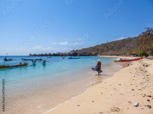 A woman in maxi dress walking on the Pink Beach in Lombook, Indonesia. Beautiful beach. Bay full of boats. No other people. Beautiful colors of the sand and the water. Dried trees in the background.