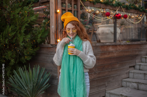 X mas time, red hair girl walking in park on a cold day, enjoying cup of hot chocolate or cocoa, wearing warm in head wear, yellow hat, knitted sweater against the decorated lights © Вероника Зеленина
