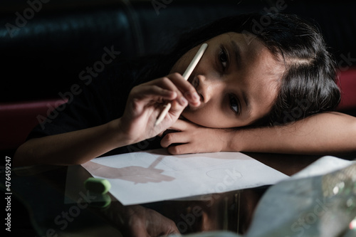 Little Asian girl with long hair is writing in pencil on a table at home, on a dark background. Kid doing homework.