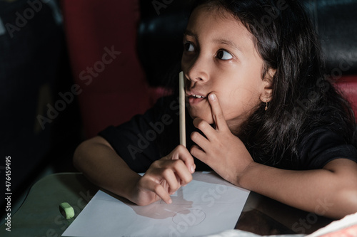Little Asian girl with long hair is writing in pencil on a table at home, on a dark background. Kid doing homework.