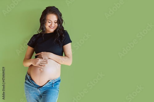 Happy pregnant woman looks at her belly and hugs it on a green background, place for text.