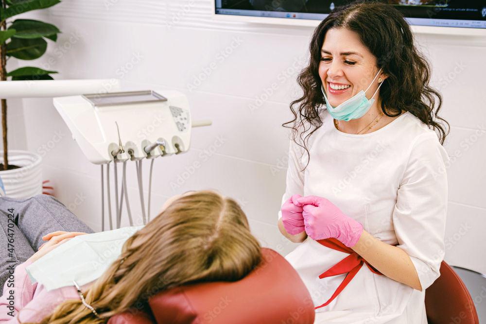 Beautiful dentist young woman smiling while wearing surgical mask for protection and gloves with a patient in the cabinet. Beautiful dentist young woman smiling while wearing surgical mask for protection and gloves with a patient in the cabinet.