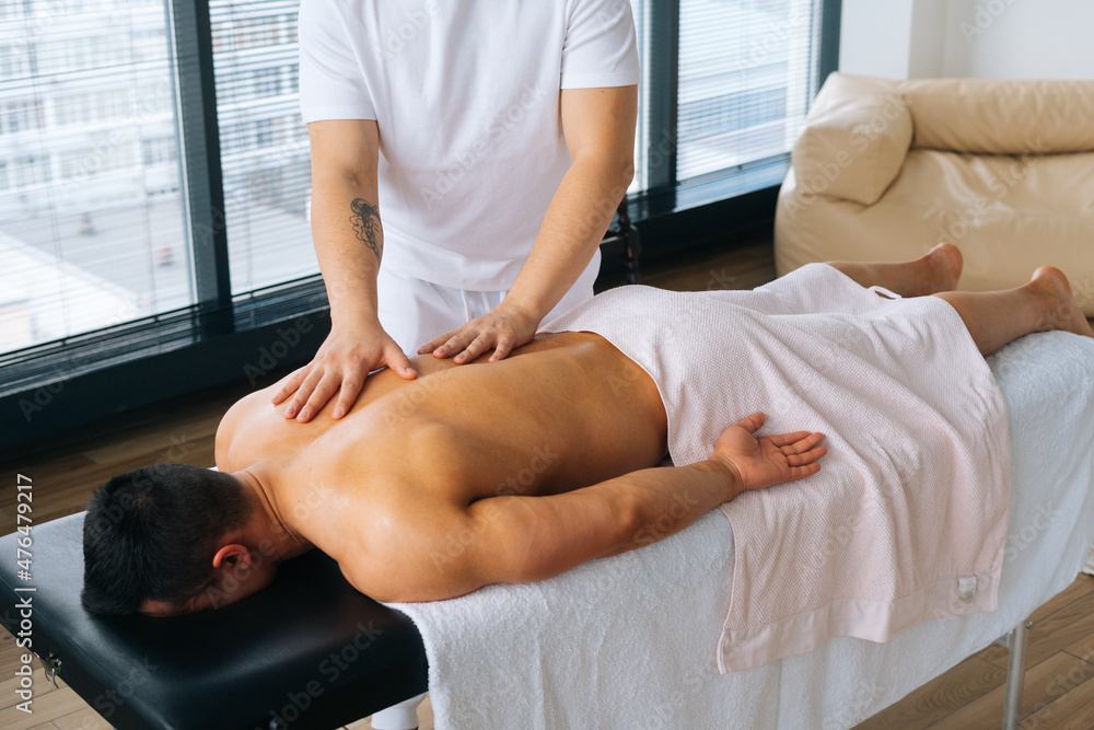 Obraz premium Medium cropped shot of professional male masseur with strong tattooed hands massaging back of unrecognizable muscular sportsman lying on stomach at massage table. Concept of healthy lifestyle and rest