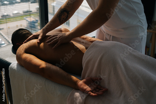 Cropped shot of unrecognizable male masseur with strong tattooed hands massaging back and shoulders of muscular sports man lying on stomach, at massage table on background of bright sunlight.