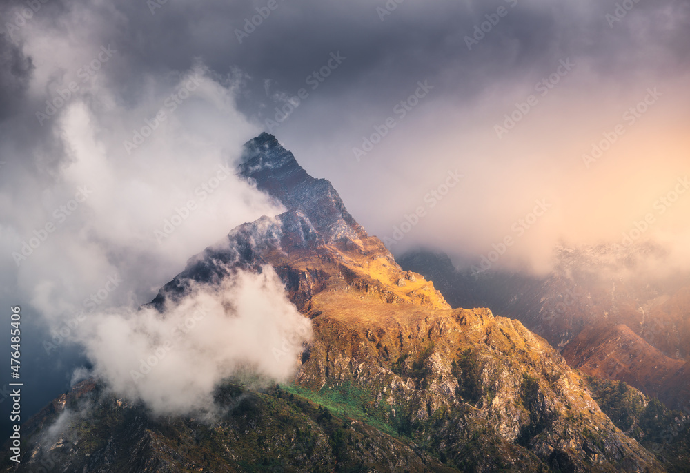 Mountain peak in low clouds at colorful sunset in Nepal. Dramatic landscape with beautiful high rocks, dramatic sky, sunlight, tress, orange grass in fog at sunset. Nature. Himalayan mountains. Travel