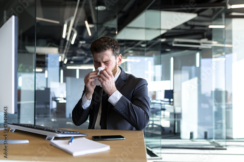 A man with a runny nose and fever works in the office, a sick businessman at the computer has a runny nose © Liubomir