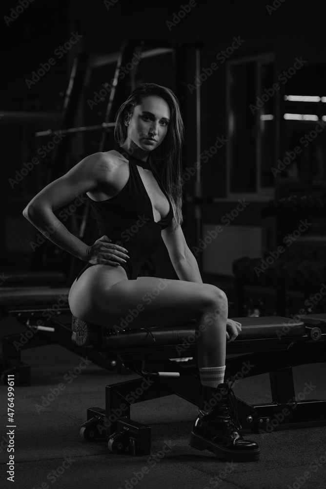 A gorgeous fit woman is posing with her arm on a waist on the bench. A muscular girl wears a black stringbody and boots after a workout in a gym.
