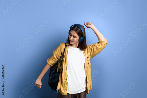 Carefree student girl is dancing, moving body and hands, looks down, smiling, enjoys life, spending free time with pleasure, wearing yellow shirt, white t-shirt, black bag and headphones.