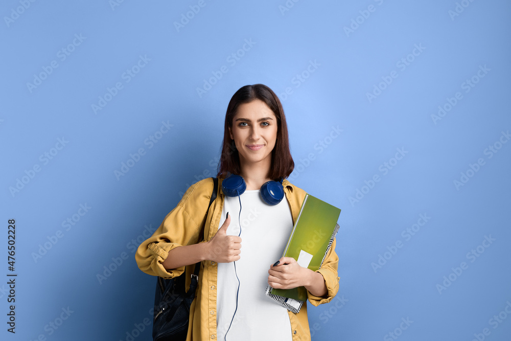 Positive smiling student girl is showing thumb up, likes an idea, supports initiative, ready to join the group, holding books, wearing yellow shirt, white t-shirt, black bag and headphones over neck. Positive smiling student girl is showing thumb up, likes an idea, supports initiative, ready to join the group, holding books, wearing yellow shirt, white t-shirt, black bag and headphones over neck.