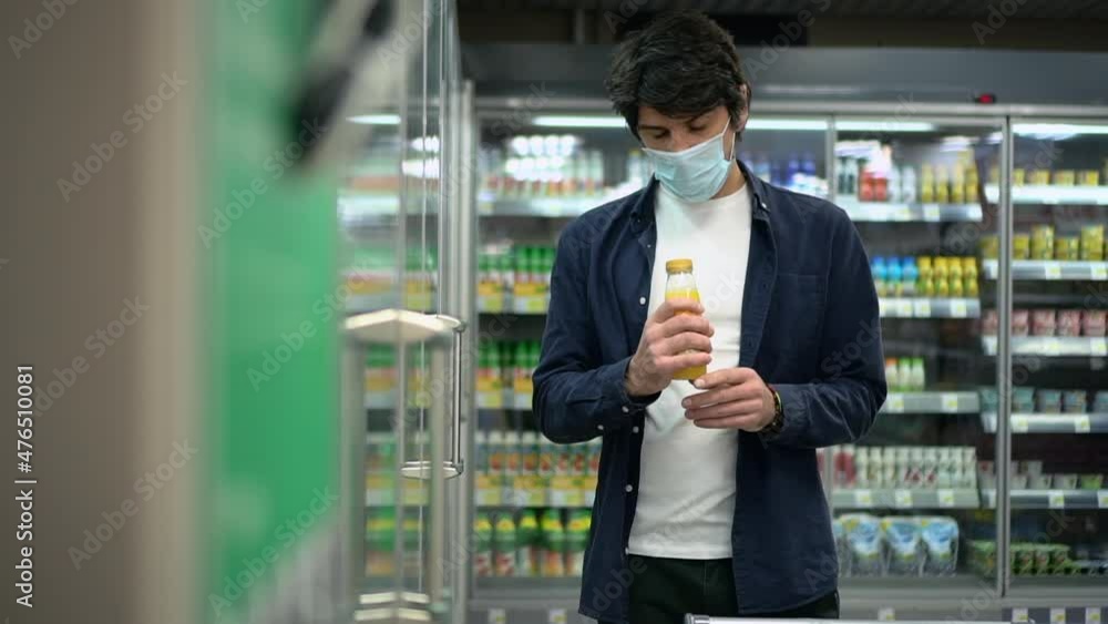 Young man choosing juice and putting in basket while shopping in store or market spbd. Caucasian male buyer wearing mask holds bottle in his hand and reads composition, makes choice and puts it in Young man choosing juice and putting in basket while shopping in store or market spbd. Caucasian male buyer wearing mask holds bottle in his hand and reads composition, makes choice and puts it in