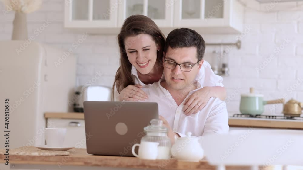 Young married people using laptop and hugging while sitting at table in apartment room spbi. Closeup view of beautiful woman, man look at computer screen and talk with smiles, hug and sit at desk in Young married people using laptop and hugging while sitting at table in apartment room spbi. Closeup view of beautiful woman, man look at computer screen and talk with smiles, hug and sit at desk in