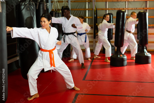 Asian woman and group of people in kimono and belts exercising with punching bags in gym during karate training.