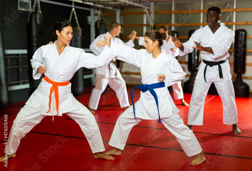 Women and man in white kimono standing in fight stance and sparring during group karate training. Their trainer, African-american man, standing beside and watching.