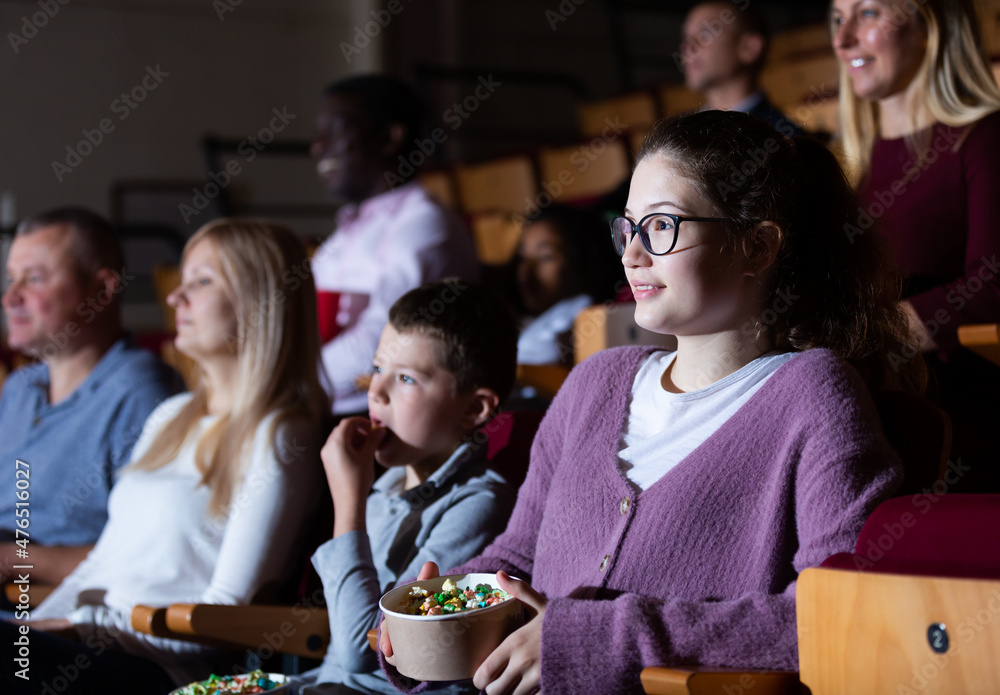 Teenage girl with her parents and preteen brother sitting in comfortable chairs in cinema, absorbedly watching movie