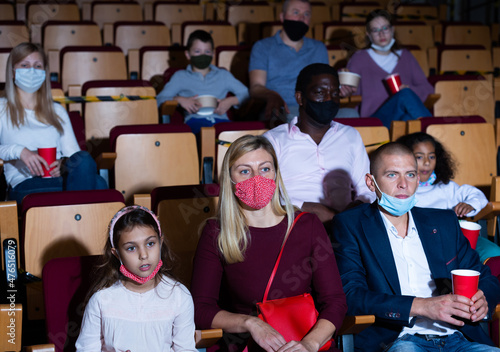 Men, women and children in protective face masks sitting in cinema auditorium. Coronavirus pandemic safety rules, social distance during movie watching © JackF