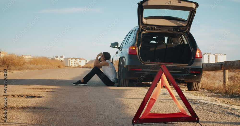 Young woman sitting a side of breakdown vehicle with flat tire using smartphone, car blinking