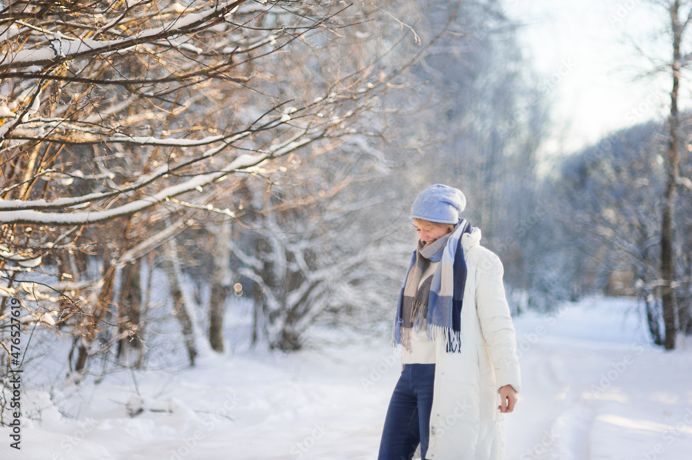 an attractive happy blonde woman in a white coat and a blue hat walks through a snow-covered park on a sunny frosty day. Healthy and active lifestyle. Place for text