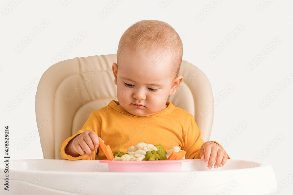 A newborn child aged one year tries boiled vegetables for the first time. A baby carefully takes a boiled carrot while sitting on a feeding chair on a white background A newborn child aged one year tries boiled vegetables for the first time. A baby carefully takes a boiled carrot while sitting on a feeding chair on a white background