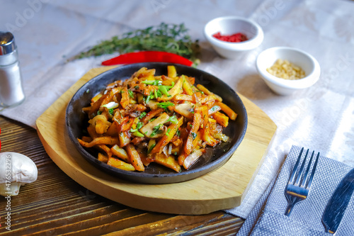 In a frying pan, fried potatoes frame pieces of meat stewed in sauce, cutlery, red bell peppers, champignons, sauce, hot peppers, spices, rosemary branch are on the table.