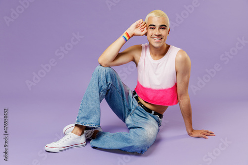 Full body happy young latin gay man 20s with make up wearing bright pink top sitting looking camera isolated on plain pastel purple background studio portrait. People lifestyle fashion lgbtq concept.