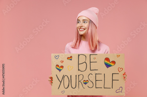 Young lesbian woman with bright dyed rose hair in rosy top shirt hat hold card sign with be yourself title text isolated on plain light pastel pink background studio. People lifestyle fashion concept.