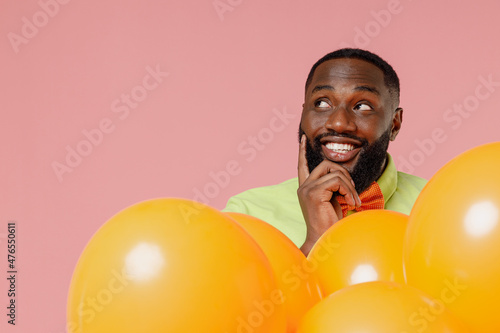 Young minded fun black gay man 20s in green t-shirt bow tie hold bunch of air inflated helium balloons celebrating birthday party look aside isolated on plain pastel pink background studio portrait © ViDi Studio
