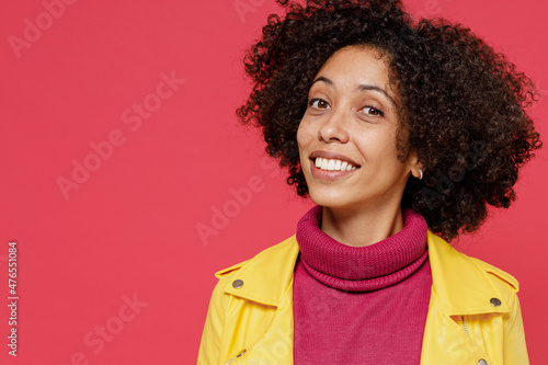 Close up bright happy ecstatic young curly black latin woman 20s wears yellow jacket chin up loking camera smiling isolated on plain red background studio portrait. People emotions lifestyle concept.